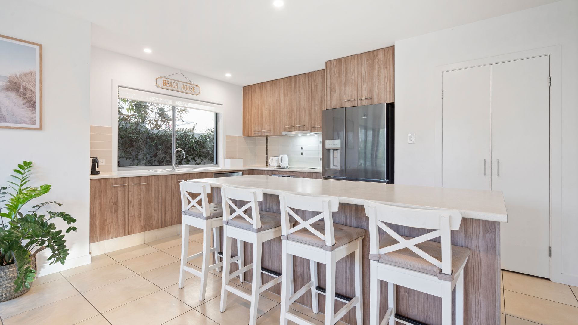 A modern kitchen with wooden cabinets, a large island with bar stools, a refrigerator, and a window with a view of greenery.