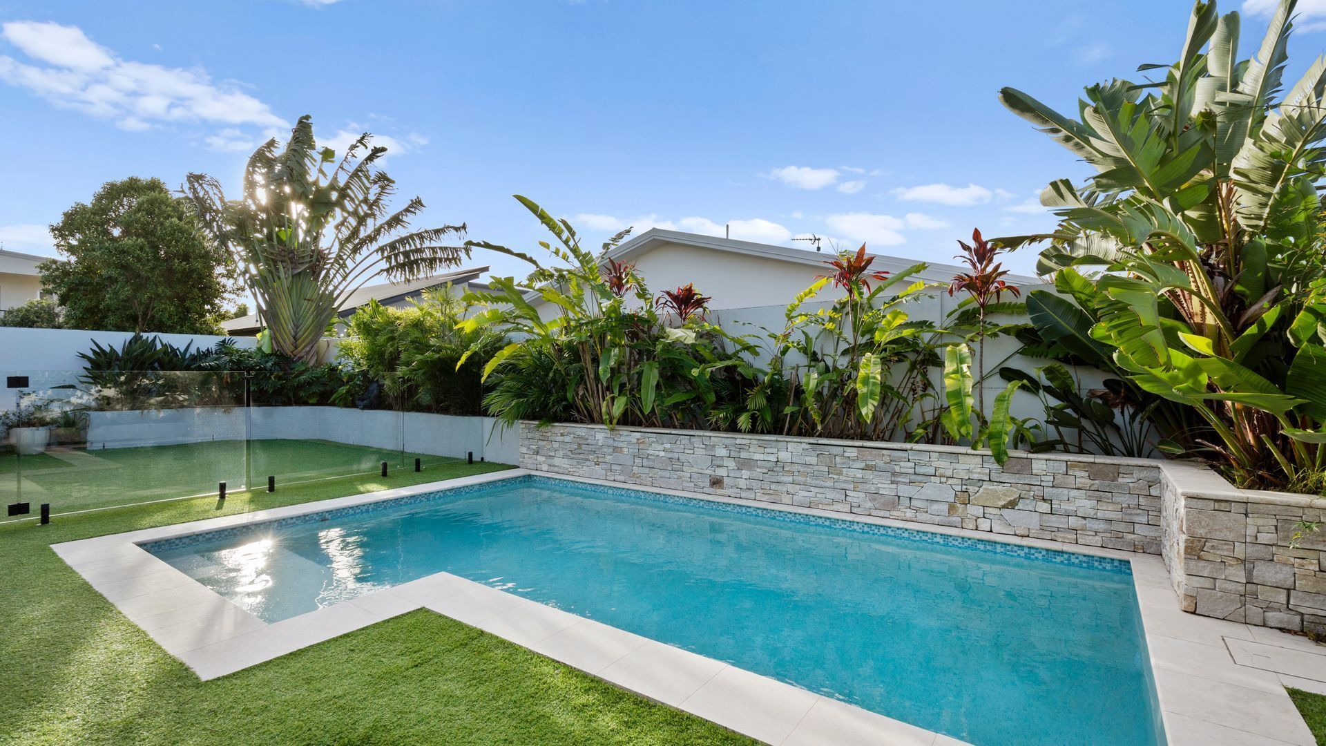 Une piscine avec un mur en pierre et une pelouse artificielle verte.