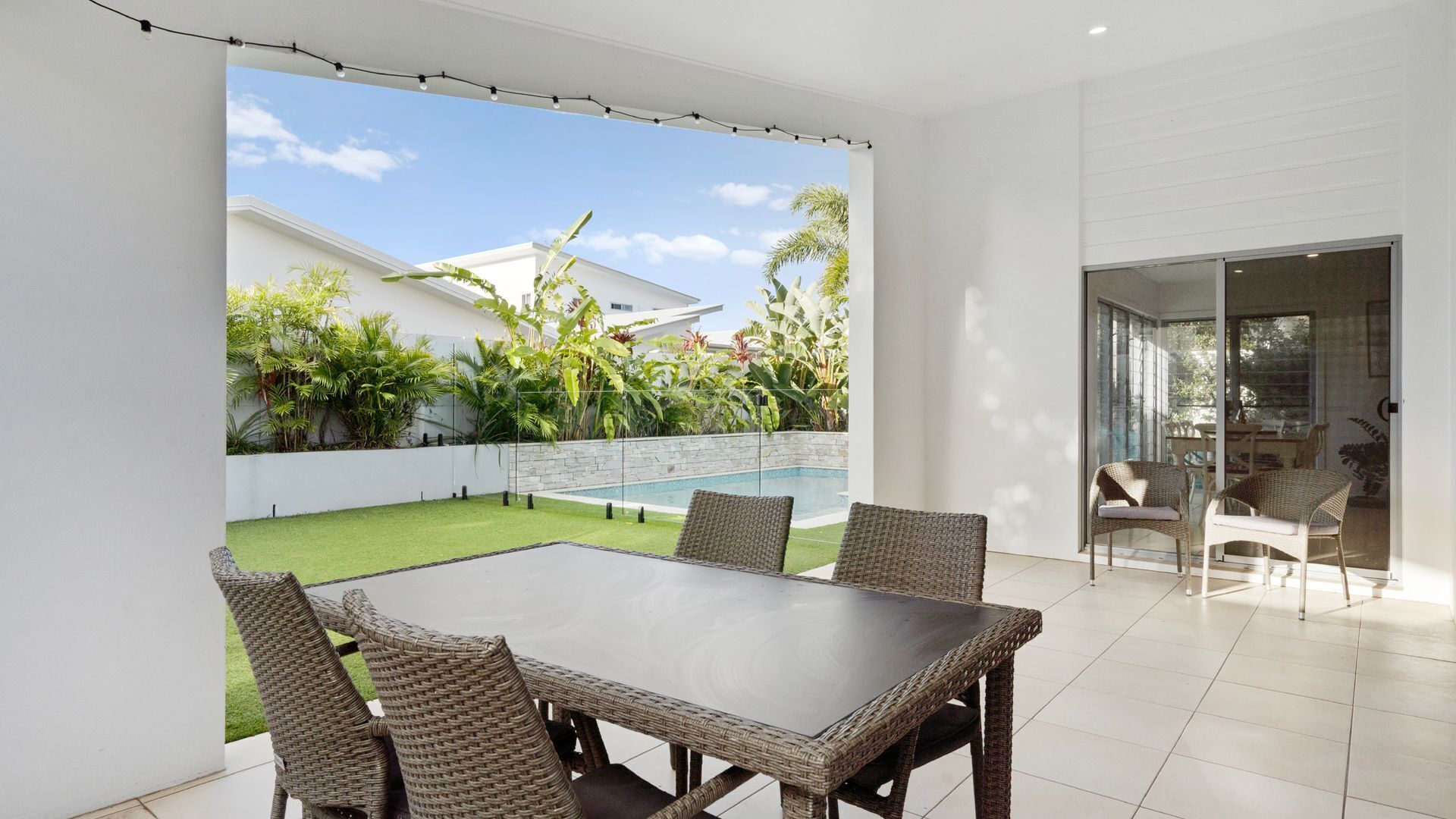 A patio with a dining table and chairs, a pool, and a view of a house and greenery.