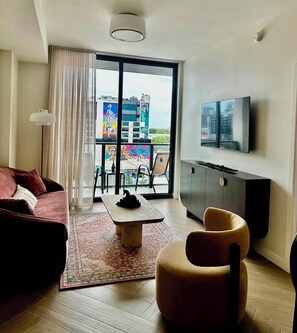 Un salon moderne avec un canapé, une table basse, un téléviseur et une vue sur le paysage urbain, grâce à de grandes fenêtres.