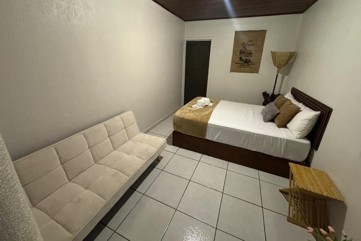 Una habitación pequeña y bien iluminada con una cama, un sofá, una puerta y una lámpara fijada a la pared.