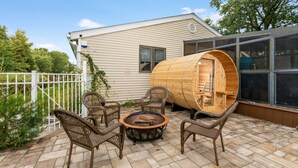 Een huis met een overdekte veranda, een vuurkorf, rieten stoelen en een houten vat.