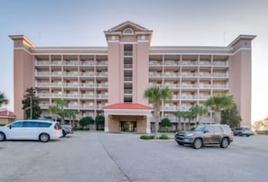 A multi-story hotel with a central entrance, balconies, and a parking lot with cars.