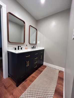 Un baño con dos espejos, un mueble de tocador oscuro, encimera blanca y una alfombra estampada.