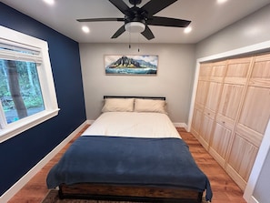 A bedroom with a bed, a ceiling fan, a window with a view of trees, and a wooden wardrobe.