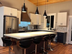 Una cocina moderna con una isla central, taburetes de bar, electrodomésticos de acero inoxidable e iluminación colgante.