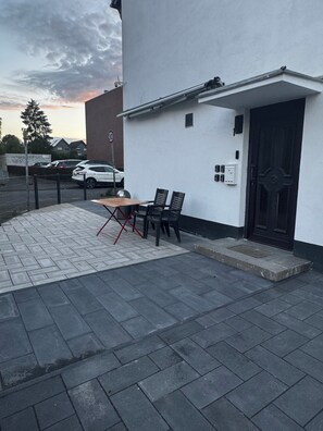 Un edificio con puerta negra, un espacio al aire libre con dos sillas y una mesa, y un patio empedrado.