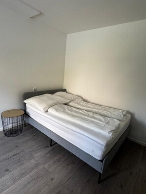 Ein Einzelbett mit grauem Gestell und weißer Bettwäsche in einem Zimmer mit Holzboden und einem kleinen runden Beistelltisch.