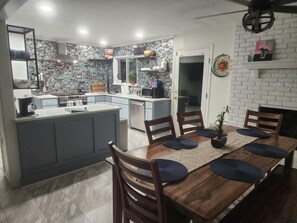 A modern kitchen with a central island, a dining area with a wooden table and chairs, and a white brick fireplace.