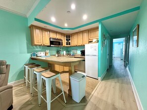 A kitchen with wooden cabinets, a central island, bar stools, a microwave, a refrigerator, and a trash bin. - Cozy Soundside Retreat at Villas of Hatteras Landing (Hatteras)