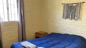A bedroom with a large bed, a window with curtains, a small wooden nightstand, and a macrame wall hanging. - Nuestras Raíces Cabins (Las Vegas)