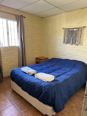 A bedroom with a large bed, a window with curtains, a small wooden nightstand, and a macrame wall hanging.