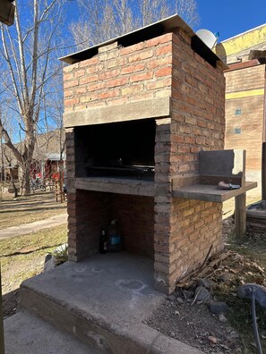 A brick-built outdoor oven with a chimney, surrounded by a grassy area and a building in the background.