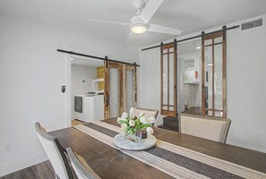 Ein Essbereich mit Holztisch, Stühlen, einer Blumenvase, einem Deckenventilator und einer Schiebetür zum Küchenbereich.