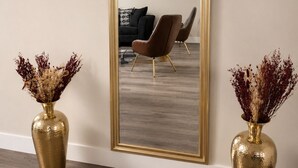 Ein Zimmer mit einem großen Spiegel, zwei goldfarbenen Vasen mit Trockenblumen, einem braunen Sofa und einem modernen Sessel.
