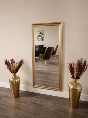 Una habitación con un espejo grande, dos jarrones dorados con flores secas, un sofá marrón y una silla moderna.