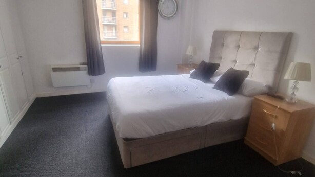 Ein Hotelzimmer mit Bett, Nachttisch, Lampe, einem Fenster mit Vorhängen, einer Wanduhr und einem Heizkörper.