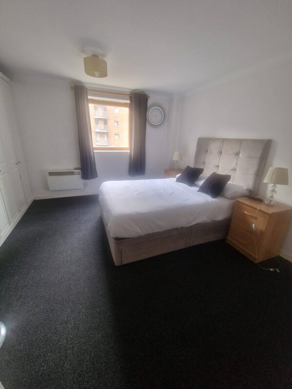 Ein Hotelzimmer mit Bett, Nachttisch, Lampe, einem Fenster mit Vorhängen, einer Wanduhr und einem Heizkörper.