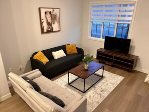 En stue med en sort sofa, en hvid sofa, et sofabord, et tv-møbel og et vindue med persienner.