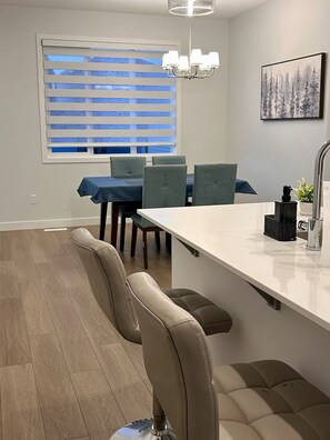 Et moderne køkken med en central ø, barstole, et spisebord med stole, et vindue med persienner og en lysekrone.