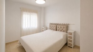 A bedroom with a bed, a nightstand, a dresser, a window with sheer curtains, and a ceiling light.