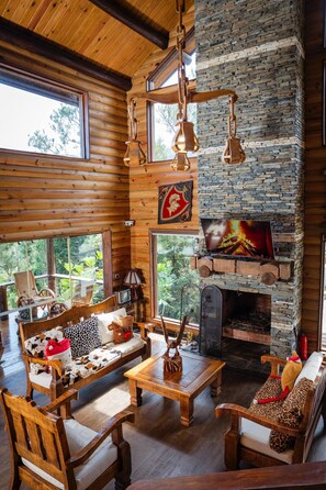 A cozy cabin interior with a stone fireplace, wooden furniture, and large windows. - Luxurious Cabin Retreat in Las Jarabacoa With Private Pool, EL Reloj SE Detuvo (Jarabacoa)