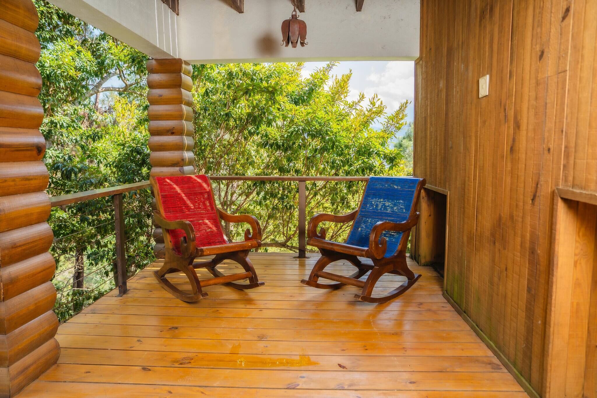 Une terrasse en bois avec deux fauteuils à bascule, l’un avec un coussin rouge et l’autre avec un coussin bleu, donnant sur un paysage verdoyant et luxuriant.
