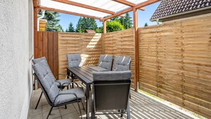 Een terras buiten met een houten tafel en stoelen, een pergola en een houten hek.
