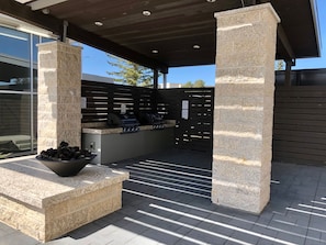An outdoor patio area with stone columns, a grill station, and a seating area with a bowl of firewood.
