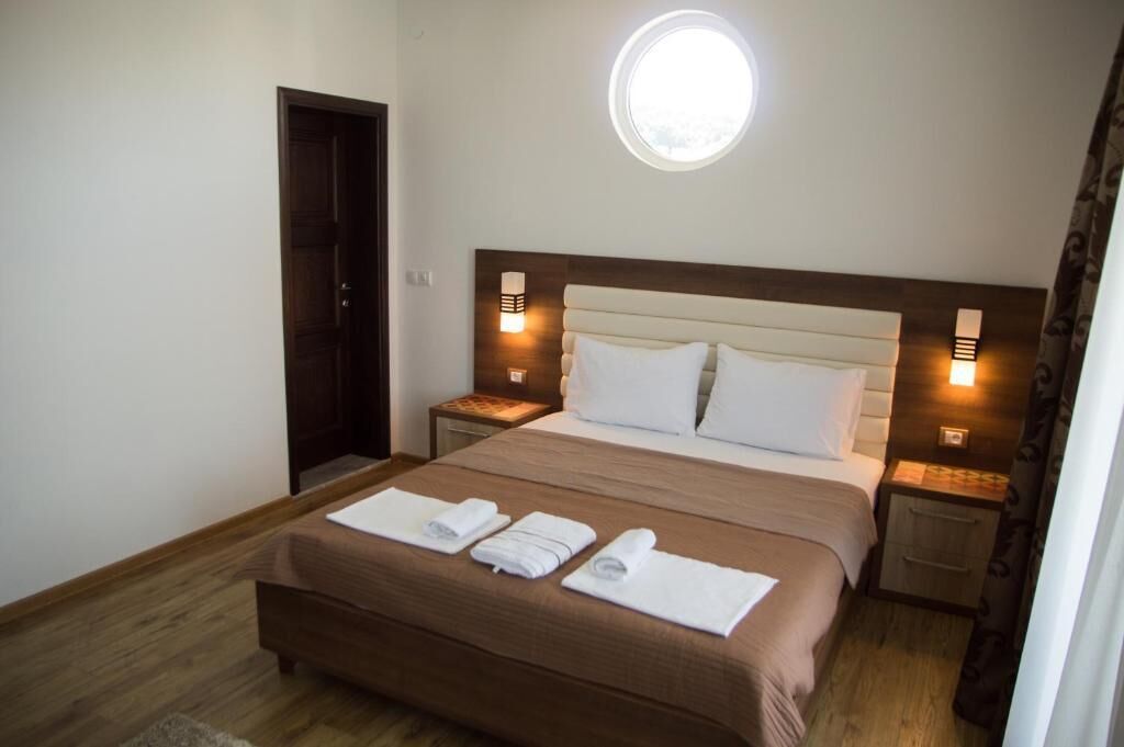 Una habitación de hotel con una cama, dos mesitas de noche, un ventanal redondo y una puerta.