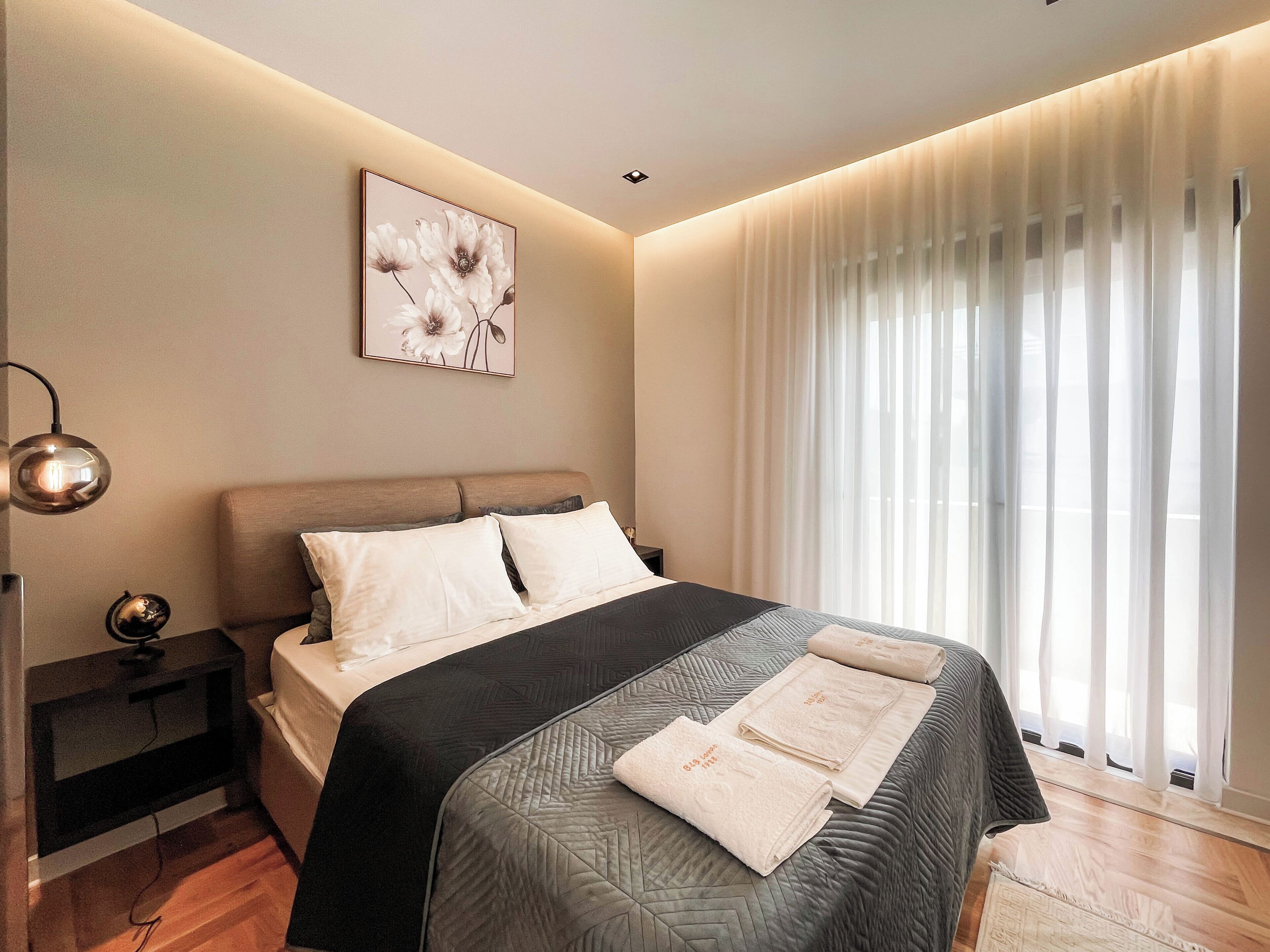 Een moderne slaapkamer met een bed, nachtkastjes, een wandlamp en een raam met dunne gordijnen.