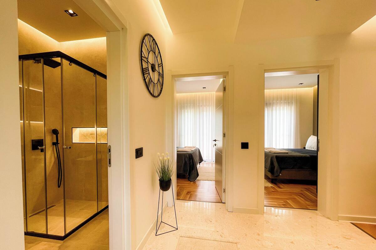 A hotel hallway with a glass shower enclosure, a large wall clock, and two bedrooms with beds and curtains.