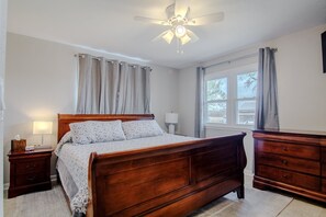 Un dormitorio con una cama de madera, una mesita de noche, un aparador, un ventilador de techo y una ventana con cortinas.