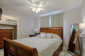 Ein Schlafzimmer mit einem hölzernen Bett, einem Deckenventilator, einem Nachttisch, einer Kommode, einem Spiegel und einem Fenster mit Vorhängen.