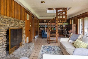 Ein gemütliches Wohnzimmer mit Kamin aus Stein, einer Holztreppe, einem Bücherregal und einem Klavier.
