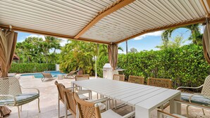 Een terras buiten met een overdekte eethoek, lounge stoelen en een zwembad.