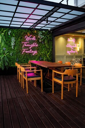Eine Holzterrasse mit Tisch und Stühlen, ein Neon-Schild mit der Aufschrift 'Words Thoughts Feelings' und eine grüne Wand mit Pflanzen.