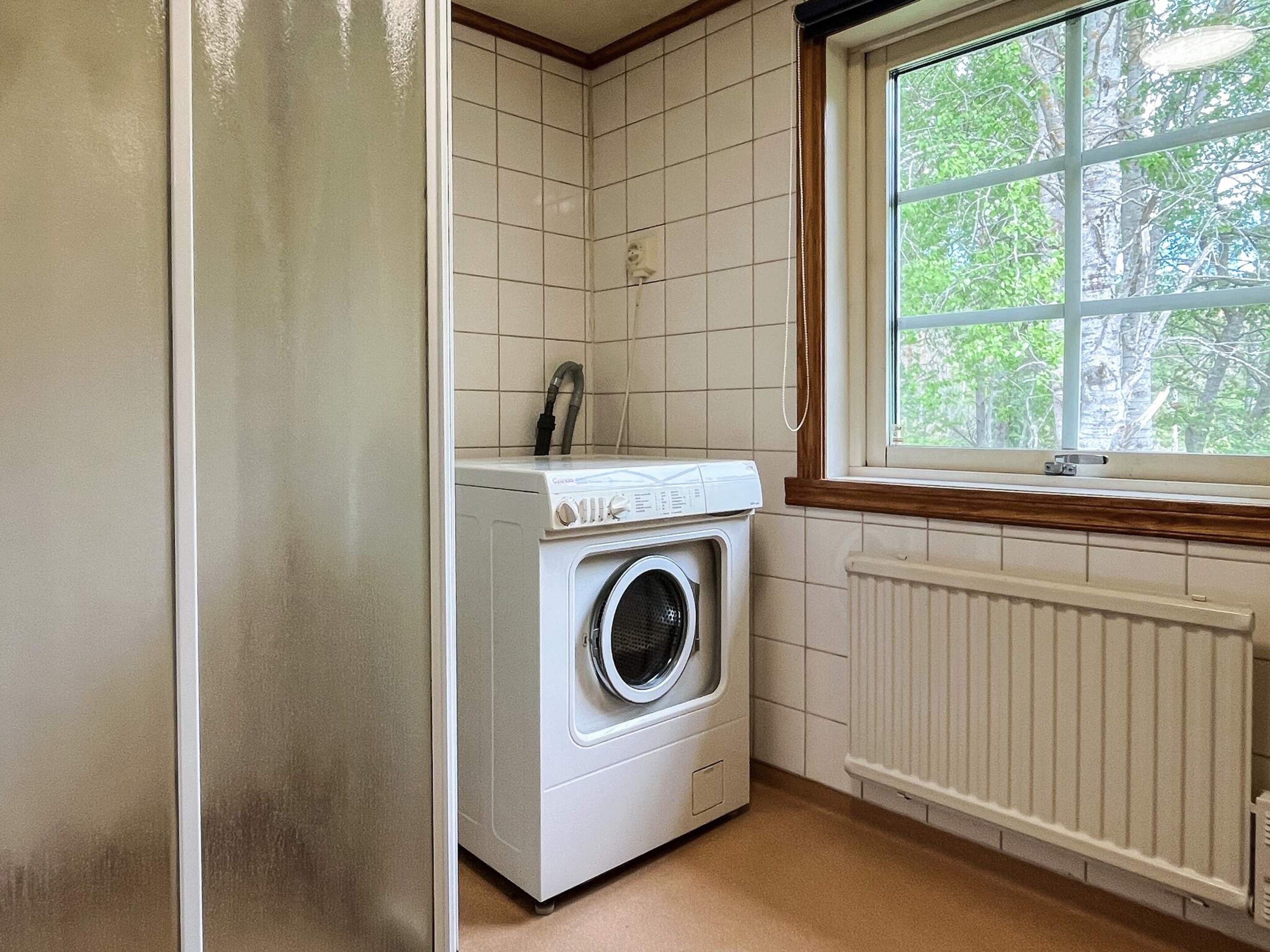 A laundry room with a front-loading washing machine and a window with a view of trees.