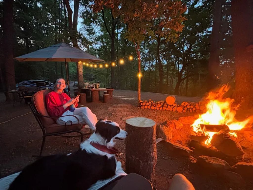 A person sitting on a bench near a fire pit, with a dog beside them.. Mountain view - Marvelous campground in Columbus perfect for your next adventure