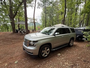 Un SUV plateado estacionado en un área boscosa con una mesa de picnic y una tienda de campaña al fondo.