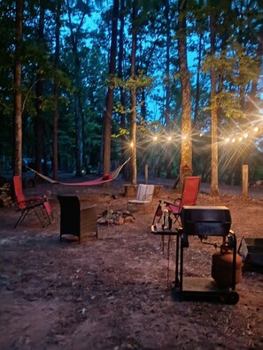A campsite with a hammock, chairs, a grill, and string lights.