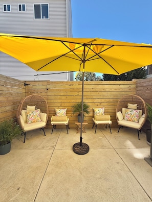 Un patio al aire libre con un paraguas amarillo, sillas de mimbre con cojines estampados y una cerca de madera.