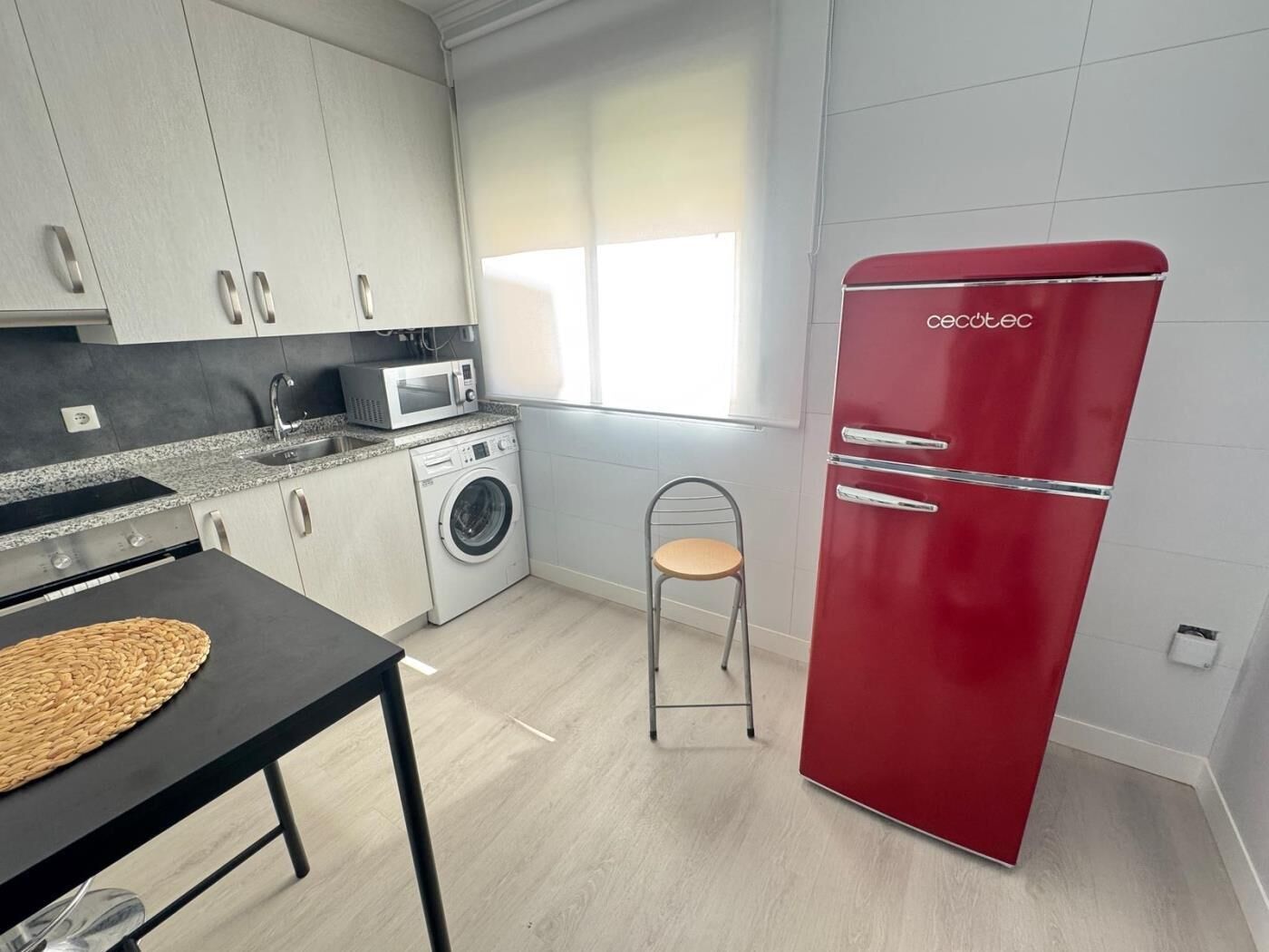 A compact kitchen with a washing machine, a microwave, a red refrigerator, a dining table with a placemat, and a window with blinds.