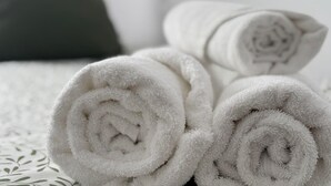 Toallas blancas enrolladas sobre una cama con una colcha estampada y un almohadón oscuro.