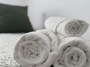 Toallas blancas enrolladas sobre una cama con una colcha estampada y un almohadón oscuro.