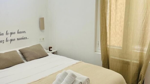 Een opgemaakt bed met kussens, een wandlamp en een raam met gordijnen.
