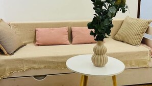 Een beige bank met twee roze kussens, een witte vaas met groene begroeiing, en een klein wit tafeltje met gouden poten.
