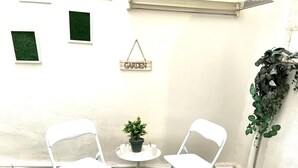Twee witte stoelen, een klein rond tafeltje, een plant in pot en een bord met de tekst 'GARDEN' op een witte muur.