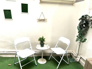 Dues cadires blanques, una taula rodona petita, una planta en test i un rètol que diu 'GARDEN' a una paret blanca.
