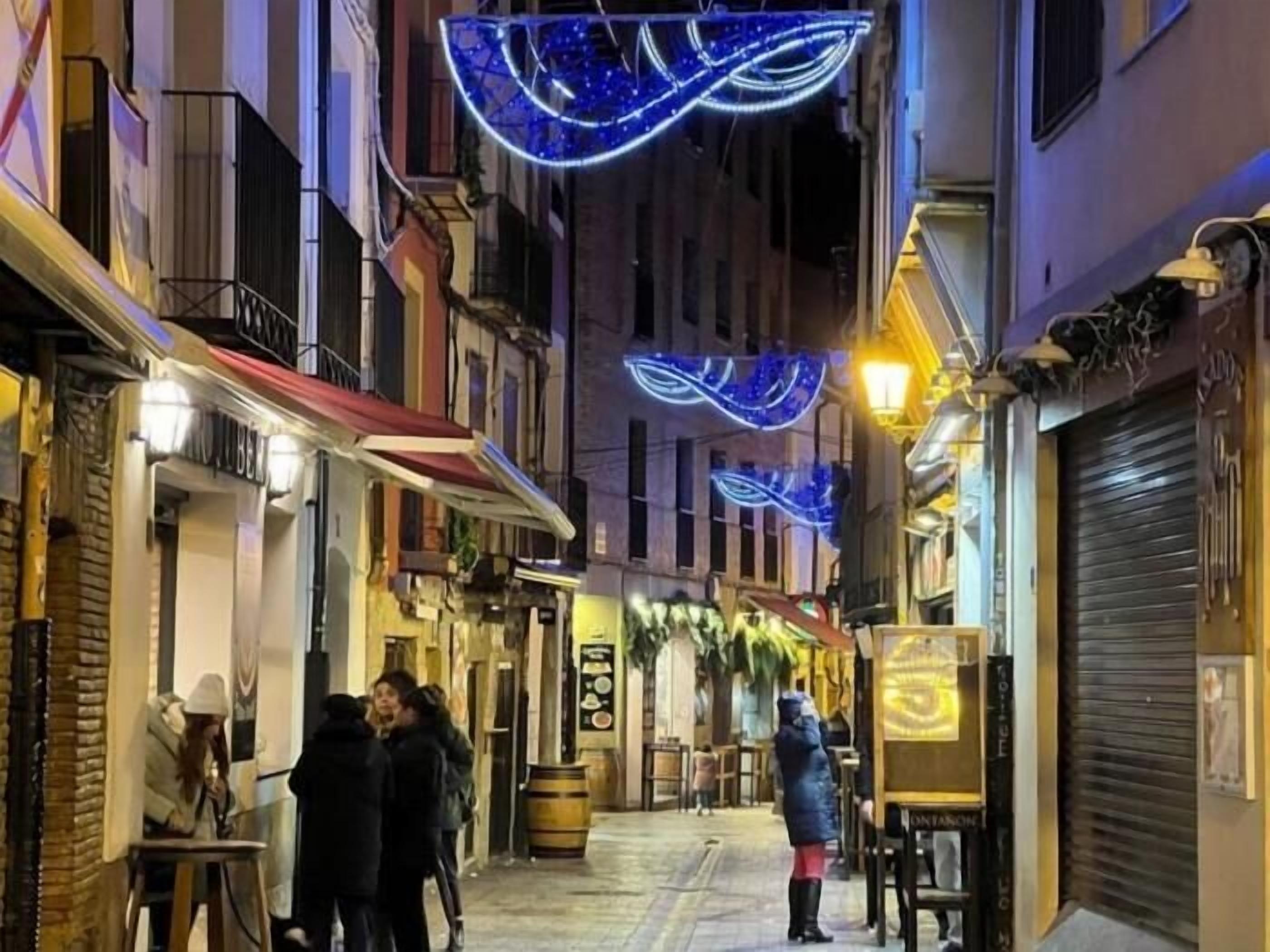 Une rue étroite, bordée de bâtiments des deux côtés, dont certains ont les volets fermés, et où quelques personnes se promènent.